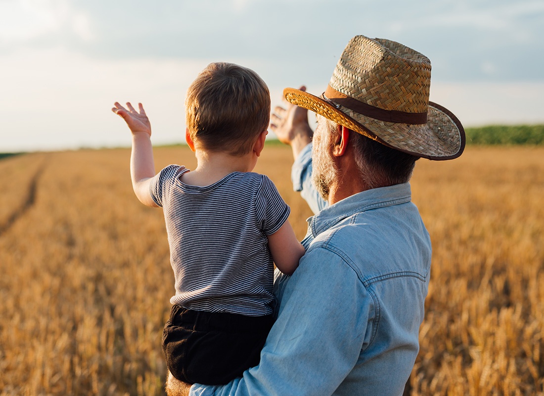 About Our Agency - Grandfather is Holding His Baby Grandson and Showing Him a Wheat Field